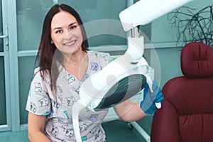 Portrait of happy female dentist sitting in the dental chair, holding lamp, smiling at the camera and ready to accept a new