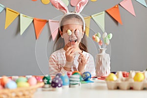 Portrait of happy funny little girl wearing rabbit ears sitting at table having fun while preparing for Easter, covering eyes with