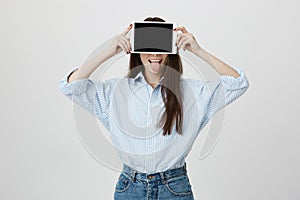 Portrait of happy and funny young european student covering face with tablet and sticking out tongue, standing over gray