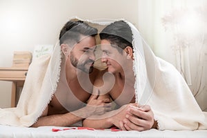 Portrait of a happy gay couple lying under a blanket on the bed together