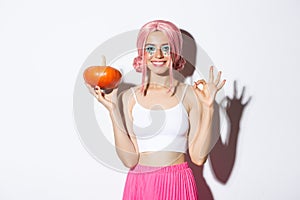 Portrait of happy girl celebrating halloween, showing small pumpkin and okay gesture, wearing cute pink wig