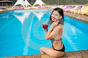 Portrait happy girl holding a cocktail, smiling and sitting on the edge of swimming pool at the resort