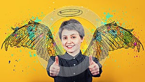 Portrait of happy girl keeps thumbs up over yellow background, smiling broadly imagining herself an angel with fluffy wings behind