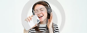 Portrait of happy girl playing karaoke app, singing into smartphone microphone, wearing wireless headphones, white