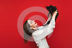 Portrait of happy girl on red background playing with cat in hands, smiling while looking at pet. Smiling girl in white shirt