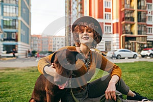 Portrait of happy girl sitting on lawn with dog, stroking puppy and smiling. Cute lady in a hat relaxing on the grass in the yard