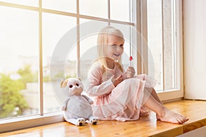 Portrait of happy girl wearing pink dress holding a lollipop while sitting on a window sill