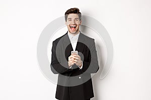 Portrait of happy good-looking man, wearing black suit, laughing from happiness and using mobile phone, standing over