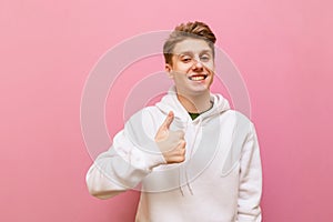 Portrait of happy guy in white casual clothes isolated on pink background, looks into the camera and shows thumb up. Smiling young