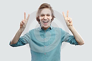 Portrait of happy handsome long haired blonde young man in blue shirt standing and looking at camera with amazing face, victory or