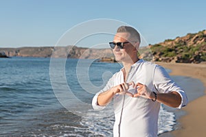 Portrait of happy handsome man in white shirt making heart gesture on the beach. Love concept