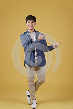 Portrait of happy handsome young asian man dressed casually pointing fingers at copyspace isolated on yellow background