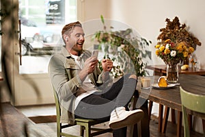 Portrait of happy handsome young man, sitting in cafe with mobile phone, wearing wireless headphones, listening to music