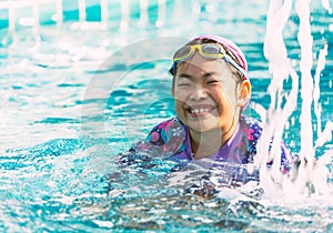 Portrait of a happy and healthy Asian child girl with goggles swimming in a swimming pool, big smiling, eyes looking at the camera