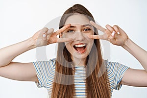 Portrait of happy and healthy girl shows peace, v-sign gesture near eyes and smiling joyful at camera, white background