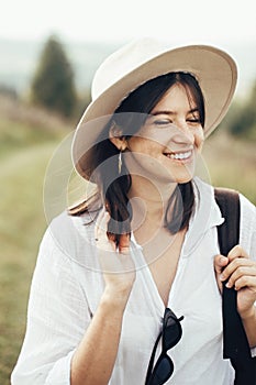 Portrait of happy hipster girl with backpack traveling on top of sunny mountain. Stylish woman in hat smiling and enjoying hiking