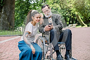 Portrait of happy hipster granddaughter with dreadlocks and barded grandfather in a wheelchair, having fun, making