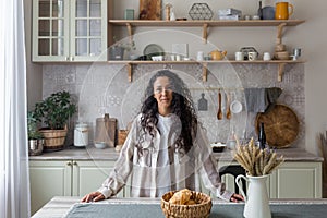 Portrait of happy hispanic woman at home in kitchen, woman with curly hair smiling and looking at camera