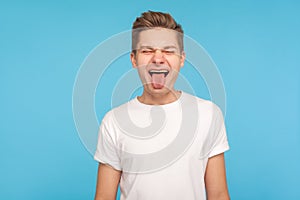 Portrait of happy humorous man in casual white t-shirt showing funny grimace and sticking out tongue