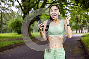 Portrait of a happy Indian woman standing in a park, holding a mobile phone in her hands and enjoying the success and