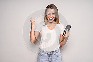 Portrait of a happy joyful girl holding mobile phone and celebrating a win over white background