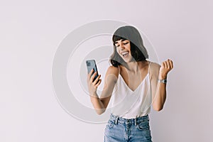 Portrait of a happy joyful girl holding mobile phone and celebrating a win over white background