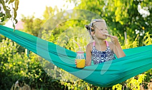 Portrait of happy kid cute girl is eating pizza and drink orange juice from jar with straw sitting on green hammock on