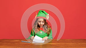 Portrait of happy kid dressed like funny gnome or elf sitting at table and writing letter to Santa Claus isolated over