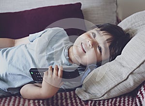 Portrait of happy kid lying on sofa watching TV, Positive child boy holding remote control looking up with smilng face. Relaxing,