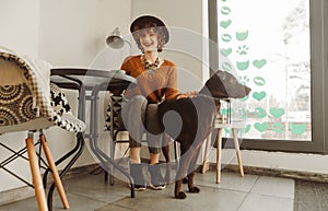 Portrait of happy lady in stylish clothes and hat sitting in chair in cozy cafe light, petting dog, looking at camera and smiling