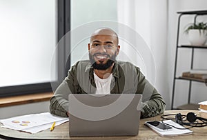 Portrait of happy latin male freelancer sitting at table, working on laptop at home office interior, copy space