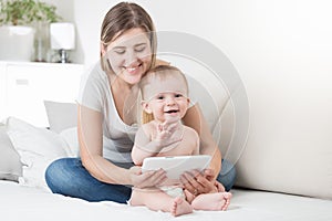 Portrait of happy laughing toddler boy and mother sitting on bed and using tablet computer