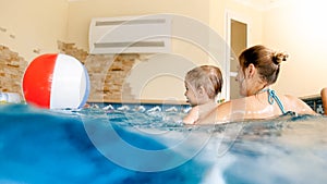 Portrait of happy laughing toddler boy with young mother playing with colorful inflatable beach ball in swimming pool at