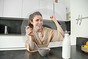 Portrait of happy, laughing young woman eating cereals with milk, triumphing, having breakfast and feeling excited