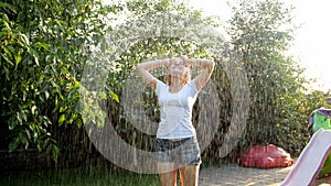 Portrait of happy laughing young woman with long hair in wet clothes dancing under warm rain in garden. Family playing