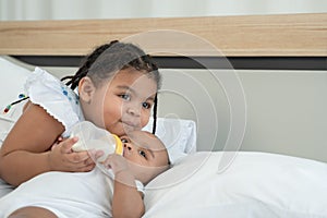 Portrait of happy little adorable African older sister feeding newborn baby milk bottle with love and care while infant lying