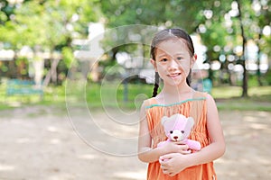 Portrait of happy little Asian child in green garden with hugging teddy bear and looking at camera. Close up smiling kid girl in