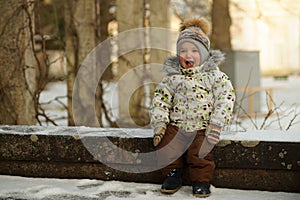 Portrait of happy little boy with red cheeks, in bright clothes and mittens sitting on bench in snowy winter park