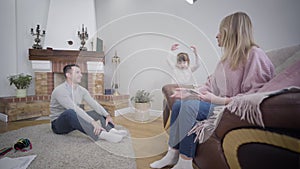 Portrait of happy little Caucasian girl showing her dancing skills to parents at home. Young married couple admiring