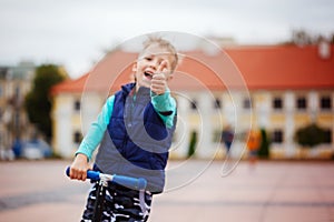 Portrait of happy little child, enjoy riding scooter on the street in the countryside, showing thumbs up. Focus on the showing th