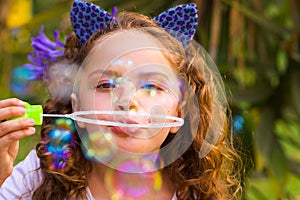 Portrait of a happy little curly girl playing with soap bubbles on a summer nature, wearing a blue ears of tiger