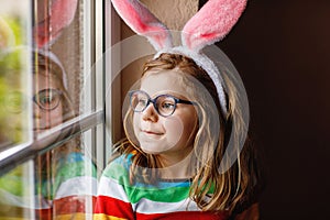 Portrait of a happy little girl with bunny ears looking outside sitting by a window. Easter holiday. Adorable child