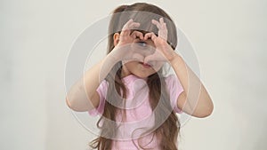 A portrait happy little girl child with brown hair smiles and holding heart shaped her hands on eyes. Healthy eyes and