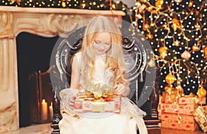 Portrait happy little girl child opens gift box near christmas tree with garland lights and fireplace at home