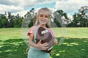 Portrait of happy little girl holding an oval brown leather rugby ball and smiling while playing with her family in park