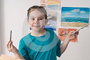 Portrait of a happy little girl painting with a brush with a palette in her hands. Little artist on the background of