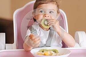 Portrait of happy little kid eating kiwi in high chair. Healthy nutrition for kids