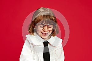 Portrait of happy little school girl in white blouse with tie and big glasses, laughing, smiling against red studio
