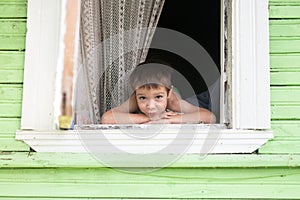 Portrait of happy little smiling boy kid is looking out of rustic wooden window and smiling. Summer time. Old rustic