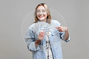 Portrait of happy lottery winner, pointing at fan of dollars, looking at camera with toothy smile. isolated on gray background